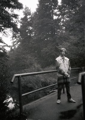Black-and-white portrait of a young man standing on a metal railing bridge, surrounded by dense forest foliage. His jacket, t...