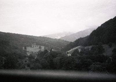 Vintage black-and-white mountain village nestled between dense forests and rolling hills, likely European alpine region. Ston...