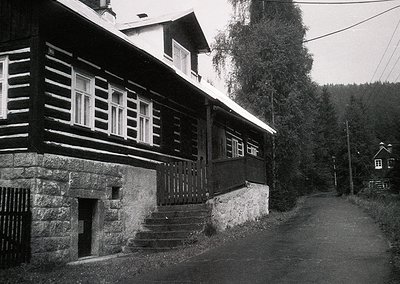 Two-story timber-framed house with stone foundation, set on a rural road. Horizontal wooden beams and white-framed windows co...