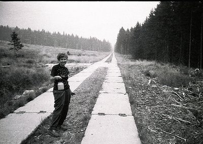 Child in 1960s-70s outdoor setting, holding a small container, standing on a paved path flanked by dense forest. Mid-century ...