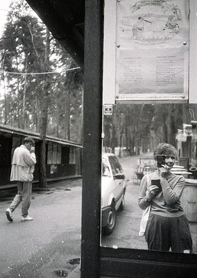 Reflection of a 1970s-era street scene in a shop window: a man in a light jacket walks past parked cars, while a woman in a s...
