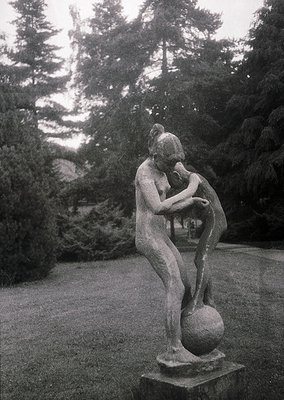 Classic bronze sculpture of a seated woman cradling a child, set in a serene garden. The composition evokes maternal tenderne...