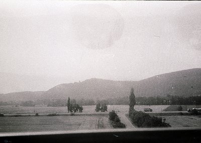Vintage black-and-white landscape featuring rolling hills, sparse trees, and a lone road winding through open fields. Overcas...