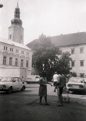 1960s-era street scene in a European town square with classic architecture: a woman in a knee-length dress and man in rolled-...