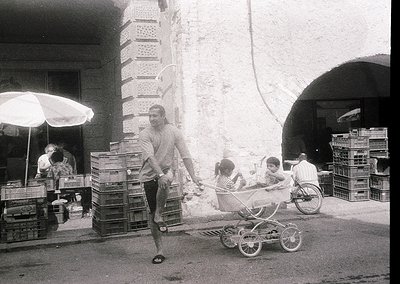 Vintage black-and-white street scene featuring a man pushing a baby stroller while balancing a crate on his shoulder. Stacked...