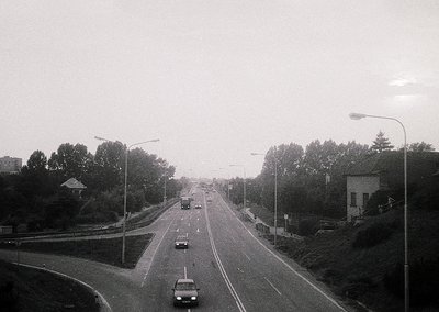 Mid-20th century urban roadway with sparse traffic, flanked by dense greenery and streetlights. Concrete barriers and a singl...