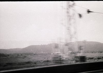 Blurred motion shot from a moving vehicle, capturing rural countryside with distant hills and sparse trees. Overhead wires an...