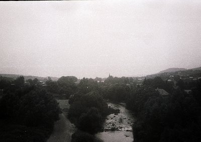 Vintage black-and-white landscape featuring a winding river flanked by dense forest. Distant village with clustered buildings...