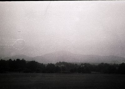 Vintage black-and-white aerial view of a flat, tree-lined landscape with faint horizon haze. Low-contrast sky shows a faint, ...