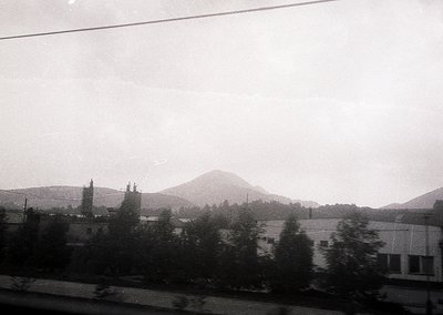 Black-and-white train window view of rural landscape with misty mountains in background. Low-lying buildings and dense greene...