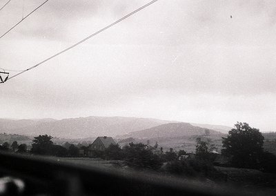 Vintage black-and-white landscape shot of rolling hills and a single-story house framed by trees. Overhead power lines and a ...