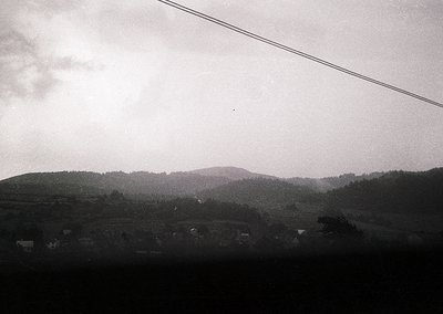 Black-and-white rural landscape featuring rolling hills, scattered farmhouses, and dense forest. Overhead power lines frame t...