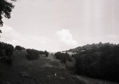 Black-and-white rural landscape featuring rolling hills, scattered trees, and a lone figure in motion near a stream. Mid-20th...