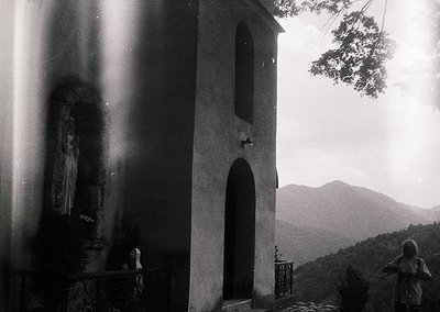 Black-and-white shot of a modest stone chapel with arched doorway, set against misty mountain backdrop. Icon of Virgin Mary i...