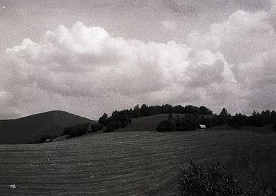 Vintage black-and-white landscape of rolling hills and open fields under dramatic cloud formations. A lone farmhouse sits nea...