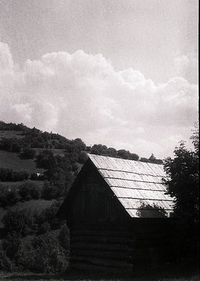 Rustic wooden cabin with gabled roof set against rolling hills and dense forest. Black-and-white monochrome evokes mid-20th c...