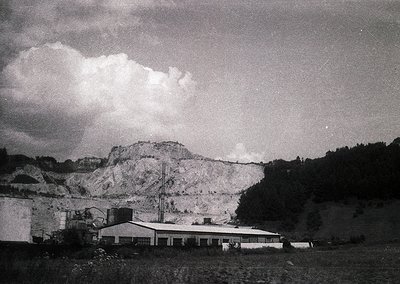 Mid-century industrial landscape featuring a low-slung, flat-roofed building beside a rocky quarry face. Dramatic cloud forma...