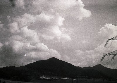 Black-and-white landscape featuring dramatic cloud formations over a forested mountain range. Power lines and utility poles f...