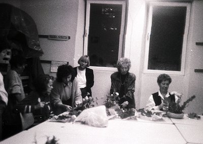 Vintage black-and-white photo of a group of women gathered around a table, crafting floral arrangements. Indoor setting with ...