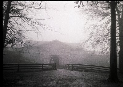 Vintage black-and-white photo of a misty pathway flanked by wooden railings, leading to a distant, partially obscured buildin...