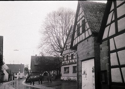 Classic timber-framed half-timbered houses dominate this mid-20th century European village street. The steep-pitched roofs an...