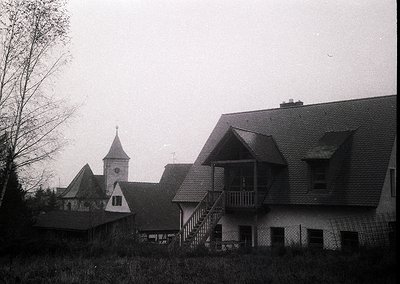 Black-and-white photo of a mid-20th century European village. Prominent gabled roofs, brick chimneys, and a church steeple wi...