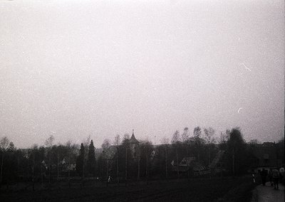 Vintage black-and-white shot of a rural cemetery with tall, uniform gravestones and a central chapel. Leafless trees frame th...