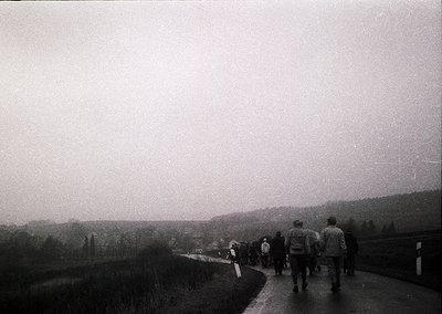 Black-and-white street scene featuring a group of 12+ pedestrians walking along a paved road flanked by grassy areas and dist...
