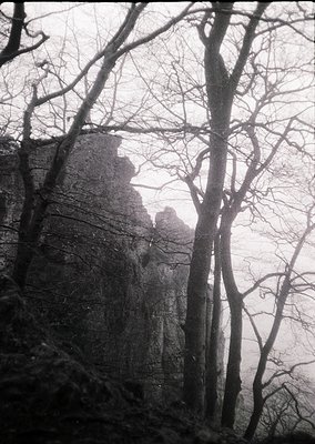 Silhouetted figures climb rocky terrain framed by bare winter trees under overcast skies. Dramatic monochrome composition evo...