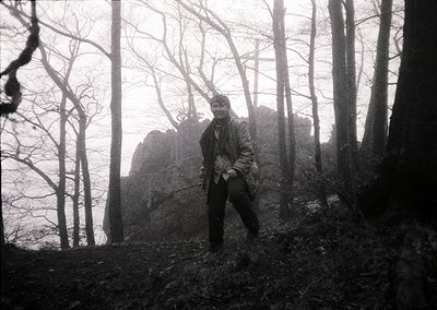 Black-and-white portrait of a lone individual in a dense forest, standing near a rocky outcrop. Clothing suggests mid-20th ce...