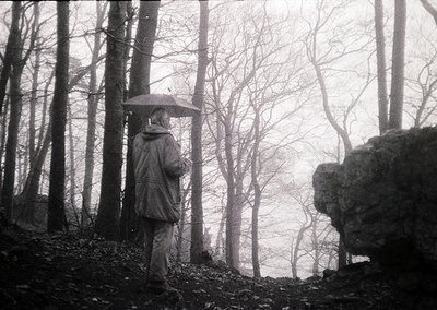 Black-and-white monochrome shot of lone figure in dense forest, holding black umbrella. Mid-century outdoor attire suggests 1...