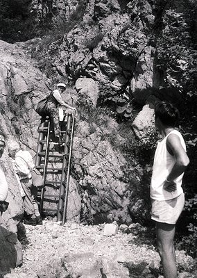 Three individuals navigate a rugged, rocky terrain using a metal ladder in mid-20th century attire. The scene suggests constr...