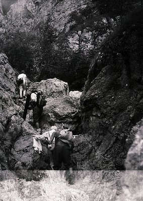 Mid-20th century black-and-white photo of three hikers navigating a rugged alpine trail with steep rock faces and misty water...