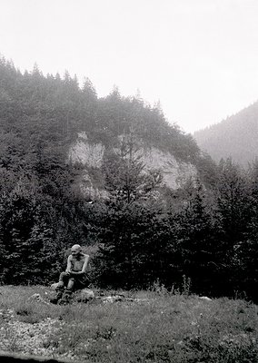 Black-and-white mountain scene featuring a lone figure in mid-hike, seated on a rock with backpack. Dense forest and misty sl...