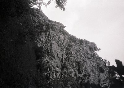 Black-and-white shot of rugged mountain terrain with dense, vertical rock face and sparse vegetation. Dramatic lighting highl...