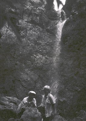 Two children stand beneath a powerful waterfall, their faces partially obscured by mist. Mid-century outdoor photography capt...