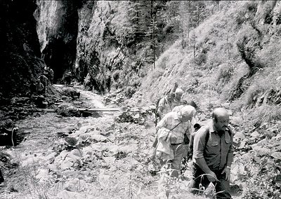 Three hikers in rugged alpine terrain, mid-20th century. Steep rock walls frame a narrow, rocky path with a trickling stream....