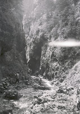 Black-and-white shot of a rugged mountain gorge with steep rock walls and a narrow, rocky path. Two hikers in mid-20th-centur...