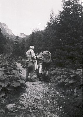 Two hikers in vintage alpine gear traverse a rocky stream crossing, surrounded by dense coniferous forest. Mid-20th century o...