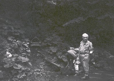 Black-and-white image of two men in rugged alpine terrain, likely mid-20th century. One man kneels near a small wooden struct...