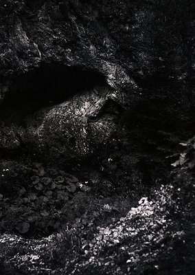 High-contrast black-and-white shot of a rugged, sunlit cave entrance with textured rock walls and floor. Light streams in fro...