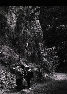 Four hikers in outdoor gear examine a rock formation in a rugged, forested area. The dramatic vertical rock face suggests a m...