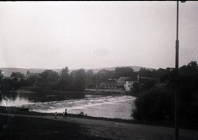 Black-and-white riverside scene with cascading waterfall, likely mid-20th century. Small group of people near water’s edge; d...
