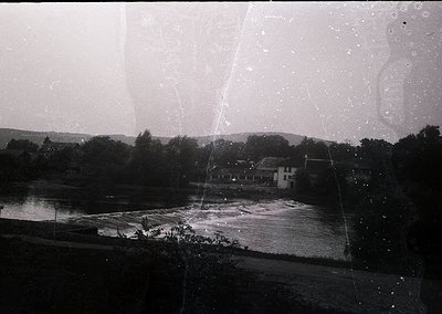 Vintage black-and-white photo of a rural riverside scene with a small cluster of buildings near water’s edge. Distinctive cur...
