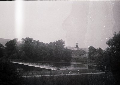 Vintage black-and-white photo of a riverside estate with classical architecture, likely mid-20th century. Prominent two-story...