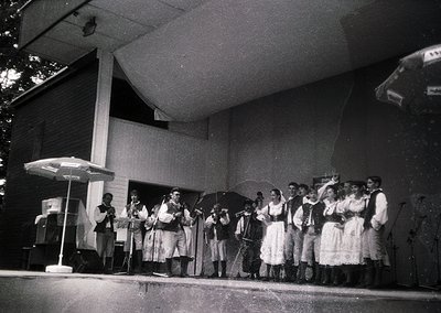 Black-and-white photo of a folk ensemble performing outdoors, likely 1960s–1980s. Traditional attire includes embroidered dre...