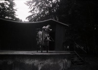 Black-and-white shot of a child on a wooden swing in a dimly lit park, framed by dense tree foliage. The swing’s metal frame ...
