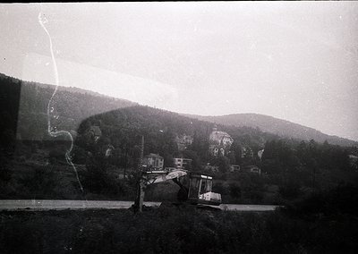 Black-and-white shot of a backhoe loader excavating on a rural roadside, framed by a window reflection. Dense forest and roll...