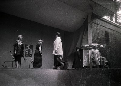 Black-and-white stage scene featuring three women in mid-20th-century attire: one playing piano, another standing beside a si...