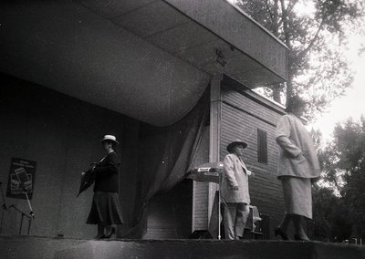 Mid-century outdoor stage performance with three musicians in 1950s-60s attire. Woman on left plays guitar; seated man at cen...